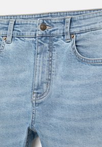 Jean en denim bleu clair en gros plan montrant la taille, le bouton avant, les passants de ceinture, la poche avant et les détails des coutures.