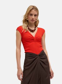 Haut en tricot rouge à côtes avec un design de devant torsadé, associé à une jupe portefeuille texturée marron. Les accessoires comprennent un collier marron épais.