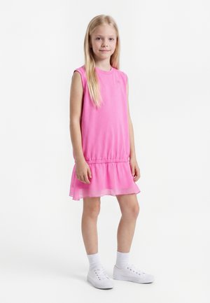 Ung pige står iført en ærmeløs lyserød kjole med flæsekant, hvide sokker og hvide sneakers på en hvid baggrund.