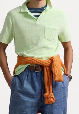 Homme portant un polo vert clair texturé sur un tee-shirt rayé, un pull orange noué à la taille, un jean bleu, une ceinture marron et une montre au poignet gauche.