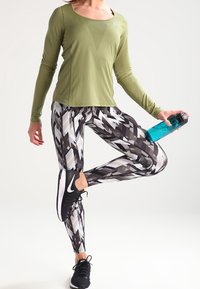 Olivgrünes Langarmoberteil aus strukturiertem Stoff, kombiniert mit Leggings mit schwarz-grauem geometrischem Muster. Hält eine blaue Wasserflasche. Schwarze Turnschuhe.