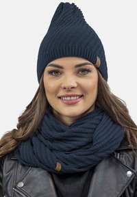 Tmavě modrý pletený kulich s špičatým vrcholem a žebrovanou strukturou, doplněný o odpovídající žebrovaný šál. Oba mají jemnou koženou etiketu.