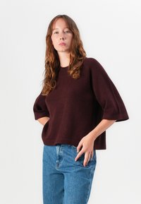Burgunderstrikkegenser med korte, vide ermer, rund halslinje og avslappet passform, kombinert med blå høyhårede jeans.