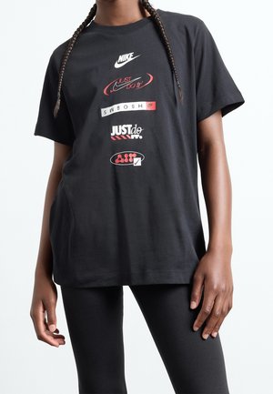 Person iført en sort Nike t-shirt med flere røde og hvide Nike-logodesign samt sorte leggings mod en hvid baggrund.