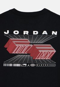 T-shirt noir avec un design graphique audacieux en rouge et blanc. Il comprend le mot "JORDAN" et un motif en grille avec des éléments architecturaux.