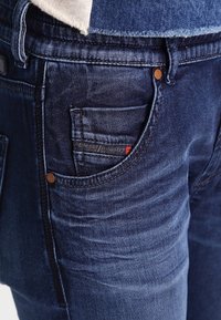 Jeans de mezclilla azul oscuro con un bolsillo trasero que presenta una etiqueta, remaches de cobre y detalles de costura visibles. Tejido de denim suave y texturado.