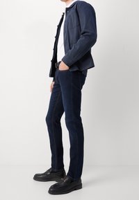 Jeans in denim blu scuro con vestibilità slim, abbinati a una camicia bianca e una giacca navy con bottoni, completati da scarpe slip-on nere.