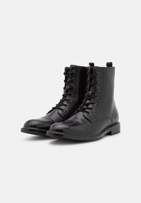 Botas de tobillo de cuero negro con diseño de cordones, puntera redonda y tacón bajo. Presentan ojales metálicos y una tira en la parte posterior.