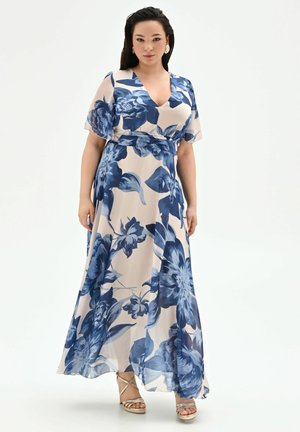 Donna che indossa un maxi abito beige con grandi stampe floreali blu, scollo a V, maniche a sbuffo e tacchi con cinturini dorati su sfondo bianco.