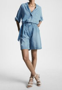 Camicia di jeans azzurro chiaro con maniche arrotolate e tasca, abbinata a pantaloni corti di jeans a vita alta e sandali bianchi con cinturini. Dettaglio con zip visibile.