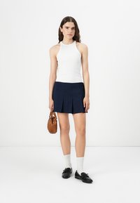 Weißes ärmelloses Oberteil mit hochgeschlossenem Kragen, kombiniert mit einem marineblauen Plisseerock. Die Accessoires umfassen eine kleine braune Handtasche und schwarze Schuhe.