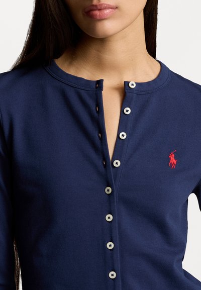 Haut en coton bleu marine à manches longues, avec un col rond, une patte de boutonnage et un petit logo rouge sur la poitrine. Texture douce et coupe ajustée.