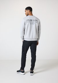 Grauer Rundhals-Pullover mit aufgedrucktem Text auf der Rückseite, kombiniert mit schwarzen Cargo-Hosen und schwarzen Sneakers. Glattes Material, lockere Passform.