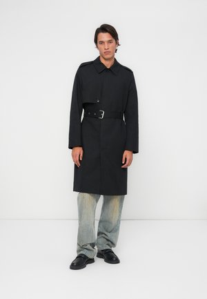 Homme adossé à un mur blanc, portant un trench-coat noir ceinturé, un jean bleu délavé et des chaussures noires, les mains détendues sur les côtés.