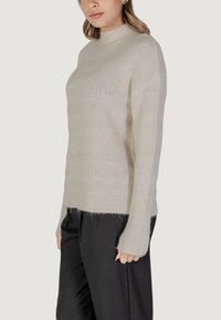 Beige, gestrickter Pullover mit hohem Kragen und langen Ärmeln. Besitzt ein strukturiertes Muster und eine leicht oversized Passform, endet an der Hüfte.