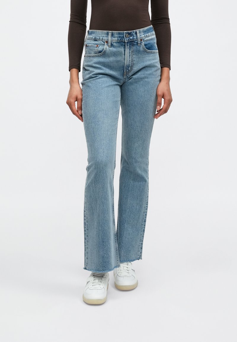 Vaqueros de mezclilla azul claro con diseño de pierna acampanada, estilo de cinco bolsillos y costuras en contraste, combinados con zapatillas blancas.