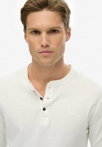 Weißes Langarm-Henley-Shirt mit drei braunen Knöpfen, rundem Halsausschnitt und strukturiertem Stoff. Kleines Logo-Detail auf der Brust.