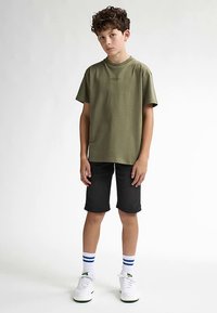 Camiseta de algodón verde oliva, cuello redondo, mangas cortas, combinada con pantalones cortos negros hasta la rodilla. Zapatillas blancas con detalles verdes y calcetines a rayas azules.