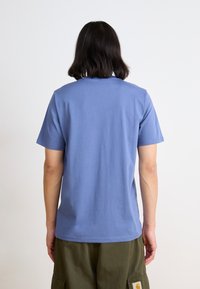 T-shirt a maniche corte in cotone azzurro chiaro, collo rotondo, texture liscia, indossato con pantaloni khaki. La vista dalla schiena mostra la semplicità nel design.