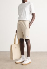 T-shirt blanc avec le logo "HUGO", short en tricot beige avec taille élastique, portant un sac fourre-tout beige clair, chaussé de sneakers blancs et beiges.