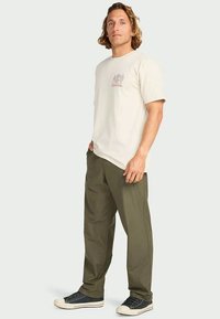 Man standing with hands in olive green pants pockets, wearing beige graphic t-shirt and black sneakers on light background.