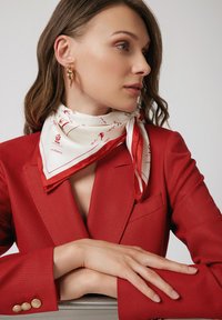 Czerwony żakiet o teksturowanej strukturze z złotymi guzikami akcentującymi. Na szyi związany jest biały szalik w czerwone wzory, który dopełnia stylizację.