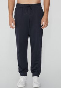 Marineblaue Jogginghosen aus weichem Stoff mit elastischem Bund, Kordelzug und gerippten Bündchen. Getragen mit weißen Sneakers.