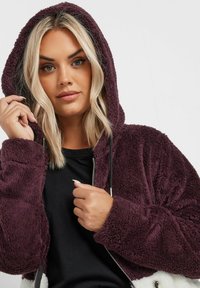 Chaqueta de forro polar en un burdeos intenso, con capucha y cierre de cremallera. Presenta un forro interior negro y una superficie exterior texturizada y suave.