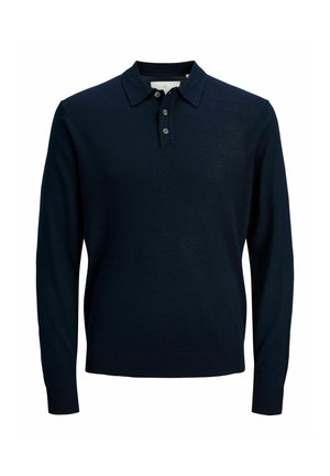 Garçon souriant debout, portant un polo marine à manches longues, un jean bleu ample et des chaussures marron sur un fond clair uni.