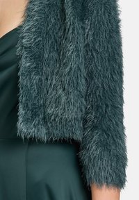 Faux-Fur-Jacke in tiefem Teal, mit einer strukturierten, strubbeligen Außenseite und verkürzten Ärmeln. Kombiniert mit einem glatten, dunkelgrünen Kleid.