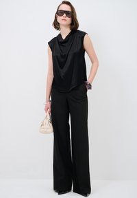 Top in satin nero con scollatura drappeggiata, abbinato a pantaloni neri a gamba larga. La modella tiene una piccola borsa beige e indossa occhiali da sole scuri.