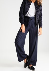 Pantaloni a gamba larga blu a righe con vita alta, abbinati a una maglietta grigia chiara e a una giacca bomber trapuntata nera. Mocassini neri.