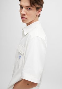 Weißes kurzärmeliges Hemd aus Baumwolle, mit einem Button-Down-Kragen, einer Tasche auf der linken Brust und einem kleinen Markenlabel-Akzent.
