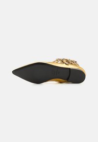 Gold flat shoe with pointed toe, embellished with metallic accents and a textured surface. Black rubber sole is visible.