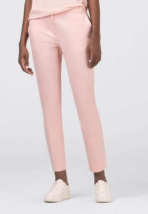 Person trägt hellrosa maßgeschneiderte Knöchelhosen, weiße Turnschuhe und ein blassrosa Oberteil, steht vor einem schlichten Hintergrund.