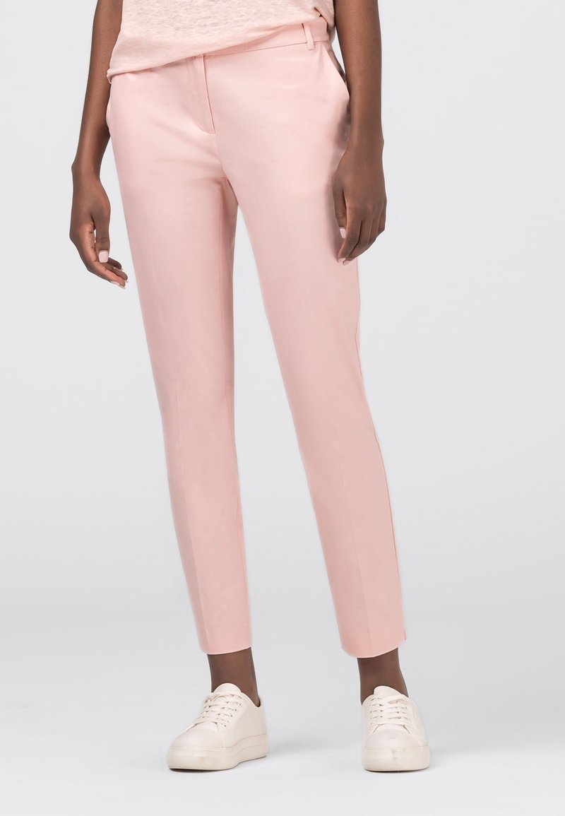 Personne portant un pantalon ajusté rose pâle coupé à la cheville, des baskets blanches et un haut rose pâle, debout devant un fond uni.