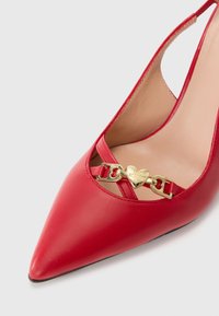 Zapato de tacón de punta afilada en cuero rojo con diseño de tirantes, herrajes dorados en forma de corazón y una correa decorativa con detalle de hebilla.