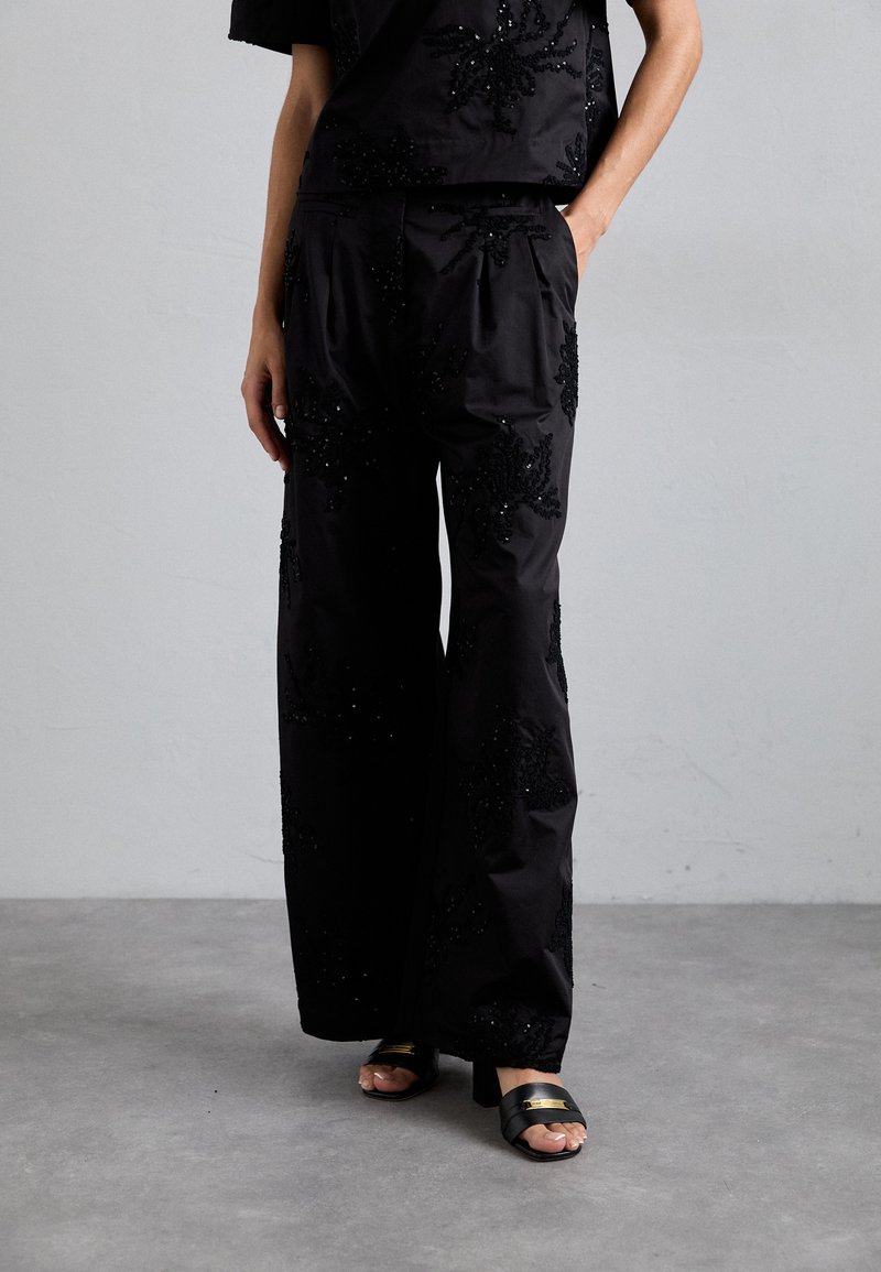 Juodos kelnės laisvu siluetu, puošnios sudėtingais karoliukais siuvinėtais gėlių raštais. Lygus, tekstūruotas audinys, platesnės kojos ir šoninės kišen�ės.