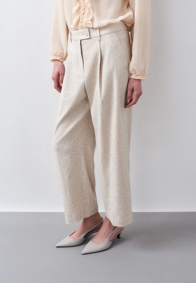 Femme portant un pantalon large taille haute de couleur blanc cassé avec un motif subtil, un chemisier beige avec des volants, et des escarpins slingbacks gris clair à bouts pointus.