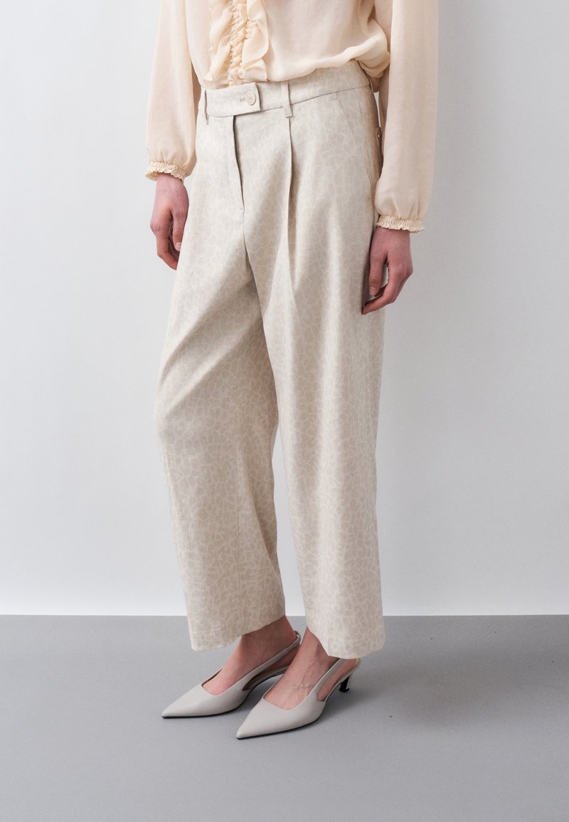 Γυναίκα που φορά ψηλόμεσο, εκρού παντελόνι με φαρδιά πόδια και διακριτικό μοτίβο, μπεζ μπλούζα με βολάν και ανοιχτό γκρι μυτερά πέδιλα με λουράκι στην πλάτη.