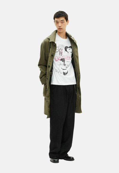 Olive green coat over a white graphic T-shirt with pink text, paired with black loose-fitting trousers and black shoes. Minimalist design.