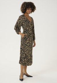 Midi haljina s uzorkom leopard u smeškastoj i crnoj boji, dugih rukava, V-izreza i džepova. Uparena sa crnim cipelama na potpeticu s otvorenom petom.