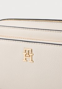 Crème leren portemonnee met een goudkleurig logo, twee ritscompartimenten en zwart accentstiksel langs de randen. Gladde textuur.