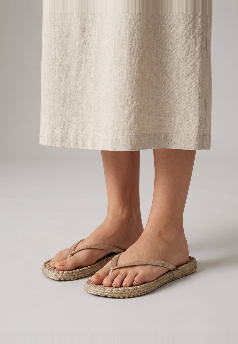 Pies con sandalias tipo chancla trenzadas beige con tiras de tela, mostrados con una falda midi texturizada de color claro sobre un fondo neutro.