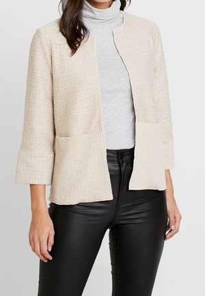 Kvinde iført en tekstureret beige jakke med åben front over en lysegrå rullekravebluse og sorte tætsiddende bukser.