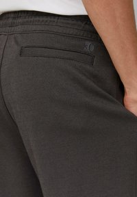 Dark gray pants feature a textured fabric, an elastic waistband, and a discreet back pocket with a small logo near the top.