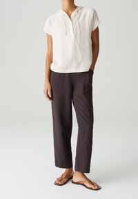 Biała lniana bluza z krótkim rękawem i sznurkiem do regulacji, w połączeniu z ciemnobrązowymi lnianymi spodniami o prostym kroju i widocznymi szwami.