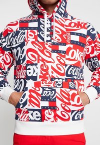 Persona vistiendo una sudadera colorida con patrones de logotipos de Coca-Cola y Tommy Jeans en rojo, blanco y azul marino, con las manos en el bolsillo frontal, combinada con pantalones rojos.