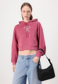 Felpa rosa corta con tasca frontale e logo bianco, abbinata a jeans blu chiaro a vita alta e una piccola borsa nera a tracolla.