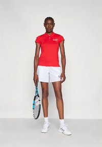 Camiseta polo vermelha com gola e mangas curtas, combinada com calções brancos. A segurar uma raquete de ténis com detalhes azuis. Design simples e desportivo.